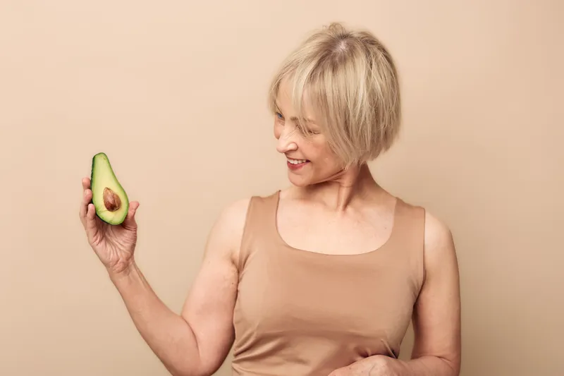 Smiling mature woman in beige top holds half of fresh avocado, looks at it with interest. Concept of healthy lifestyle, nutrition, vegan diet, and natural food choice.