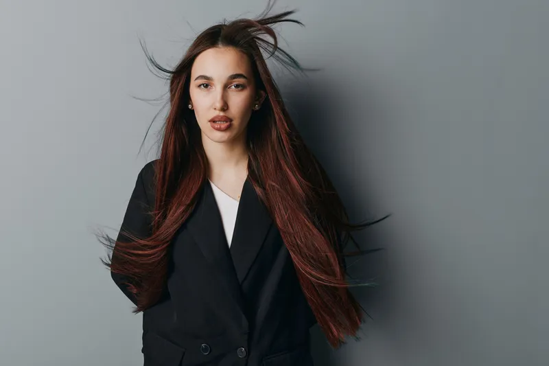 Young woman with long flowing hair wearing black blazer and white shirt posing confidently against gray background for fashion, beauty and portrait photography concept.