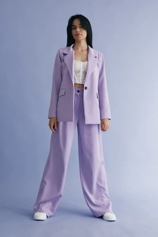 Young woman wearing light purple oversized suit and white lace top stands confidently against a pastel purple background, showcasing modern fashion style and relaxed pose.