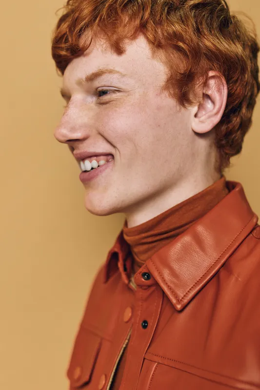 Young man with red hair smiling in profile wearing a brown leather jacket and turtleneck against a warm beige background. Casual happy expression with closed lips.