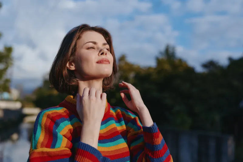 Young woman in a striped sweater with short hair outdoors, smiling and looking up by a river background. Casual outfit, mid twenties model enjoying a relaxed lifestyle moment.