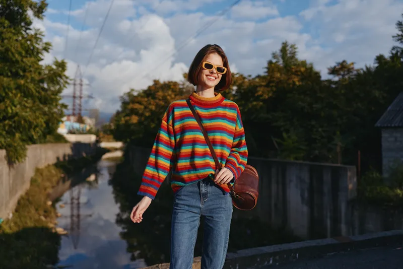 woman smiling in her mid 20s wearing a colorful striped sweater, sunglasses and jeans, walking with a brown crossbody bag by an urban canal for relaxed travel lifestyle.
