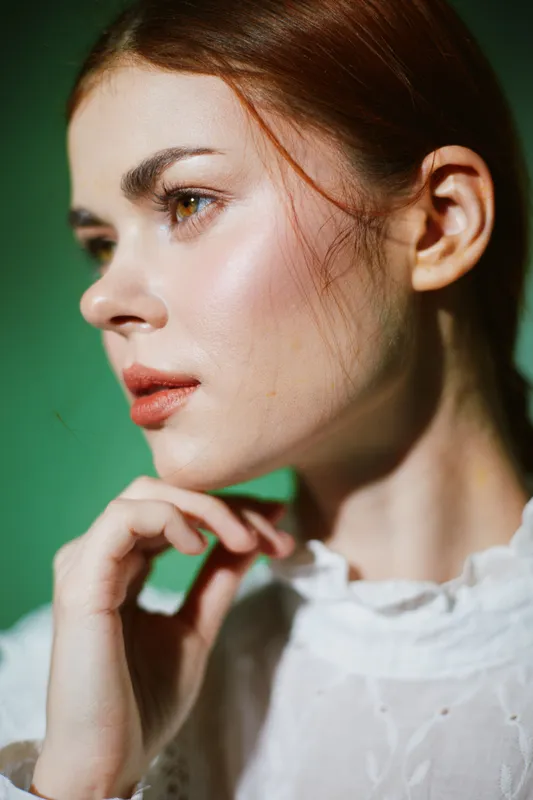 Portrait of a thoughtful young woman with red hair in a white blouse, showcasing natural beauty and gentle emotions against a green background