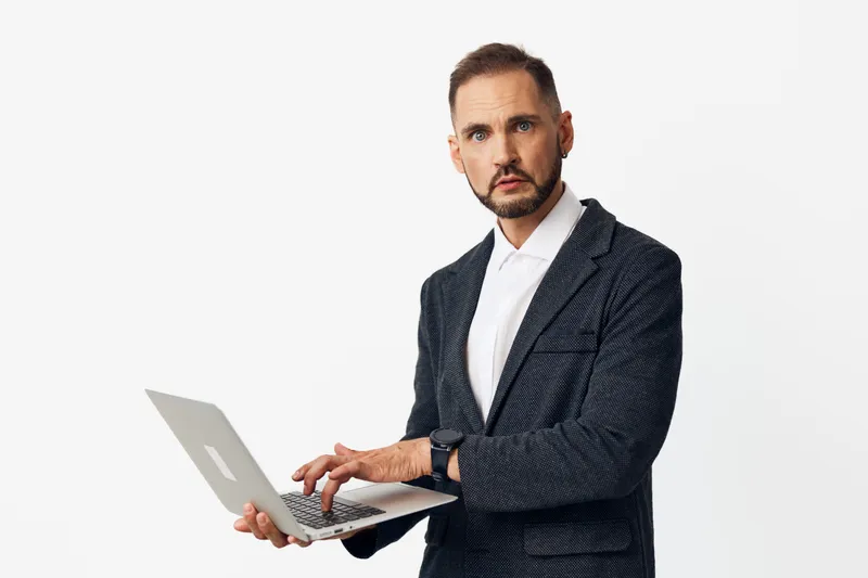 A professional male business figure stands against a solid colored background, holding a laptop and projecting focus, confidence, and readiness for work.