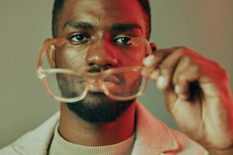 A stylish young Black man holding glasses in front of his face, showcasing a confident expression against a colorful background Fashion, emotion, and modern style combined