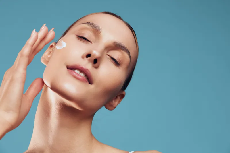 Young woman with smooth skin applies a white facial cream to her cheek in a close-up shot on a soft blue background, conveying skincare, wellness, and self care in a calm mood