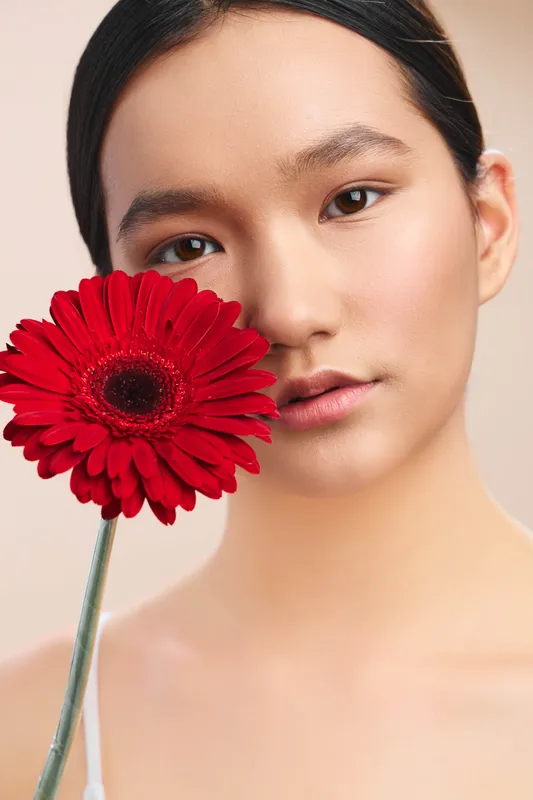 asian woman gazes softly at the camera while holding a vivid red flower near her face, creating a calm, minimalist portrait with graceful, natural light.