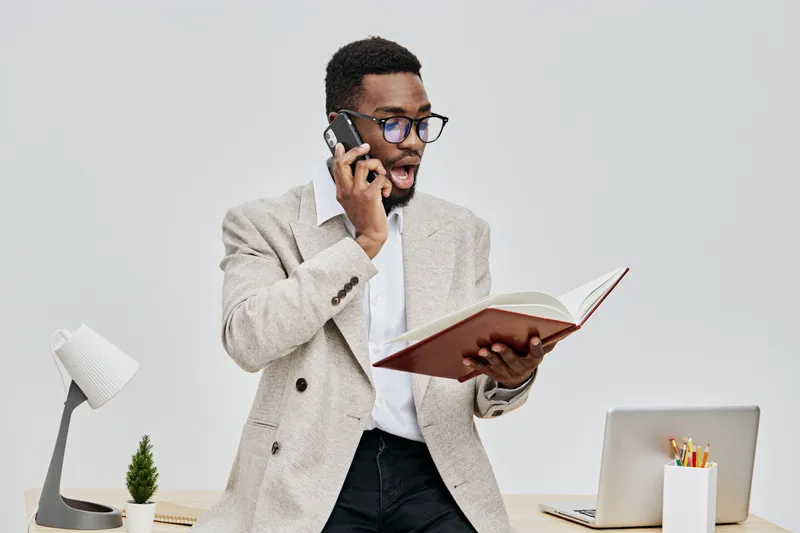 Professional businessman in trendy beige blazer and glasses talking on mobile phone while reading a notebook at a modern office workspace with white background. People lifestyle concept, communication