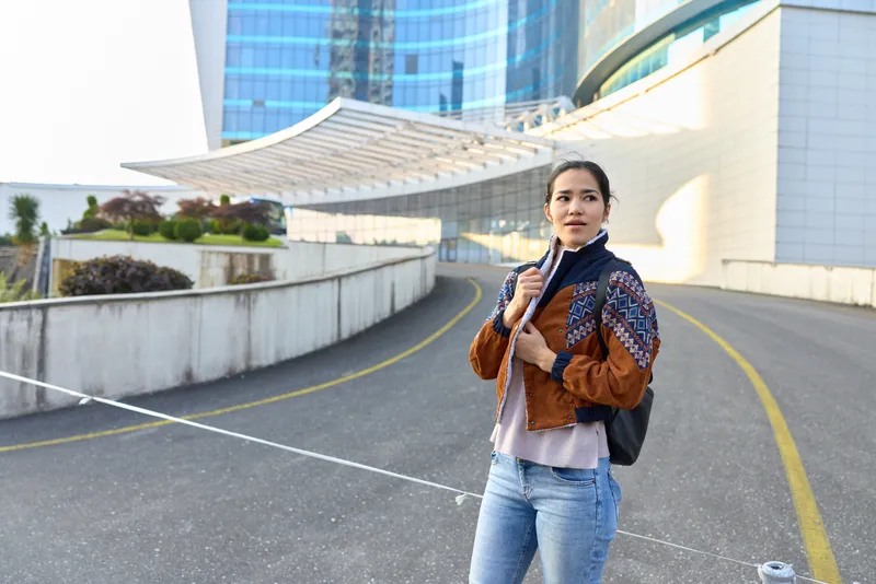 Woman in a patterned jacket with backpack stands on a wide asphalt road beside modern architecture, urban travel scene, fashionable traveler ready for a journey.