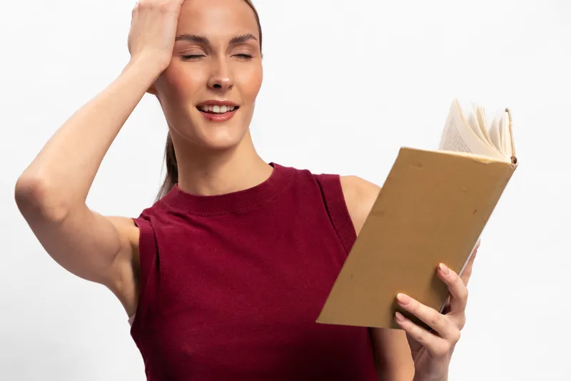 Young woman in sleeveless burgundy top holds an open book while touching her head with eyes closed. Concept for concentration, memory, and studying in bright studio setting.