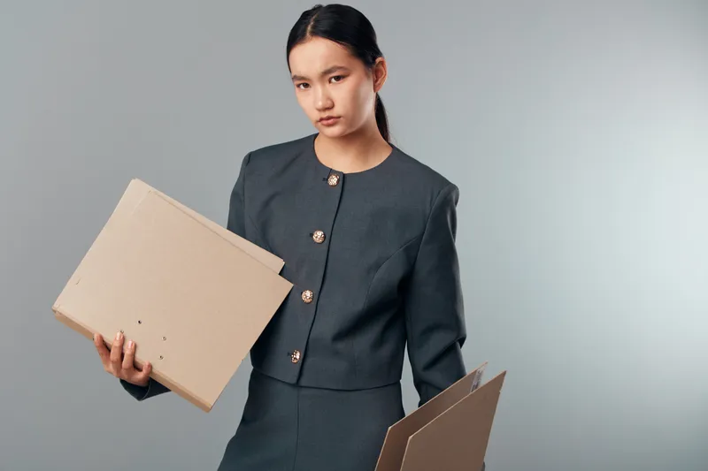 Young woman in business attire holding cardboard boxes for delivery or packaging. Female employee, logistics and shipment with document storage and order processing in studio space.