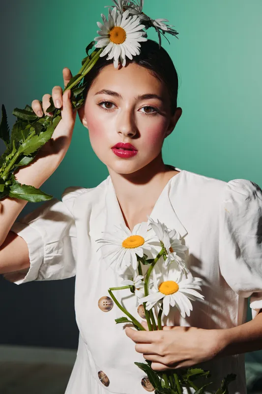 Young woman with vibrant makeup holds white daisies with yellow centers. She wears light clothing and poses against green background. Concept of beauty fashion creative minimalism.
