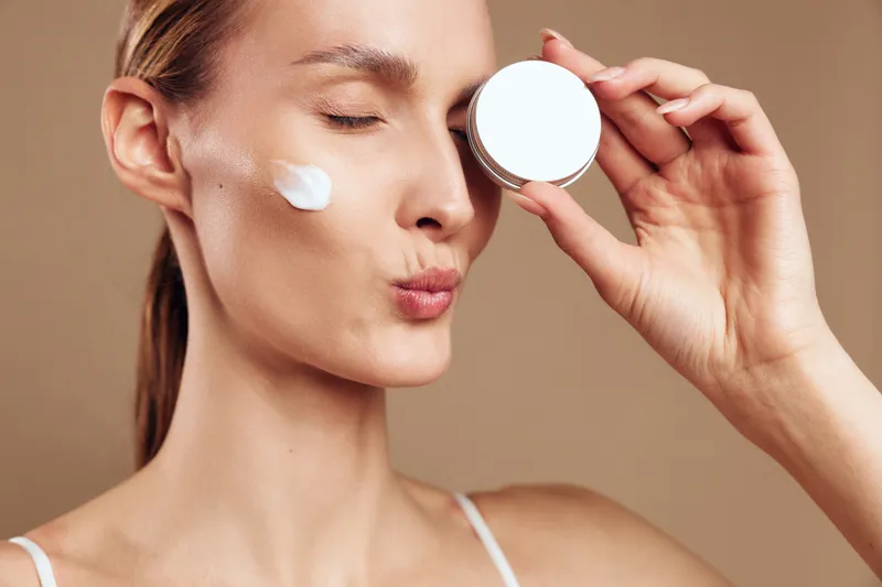 Young woman with blonde hair applies face cream on her cheek while holding a jar near her closed eyes against a beige background in a skincare beauty routine.
