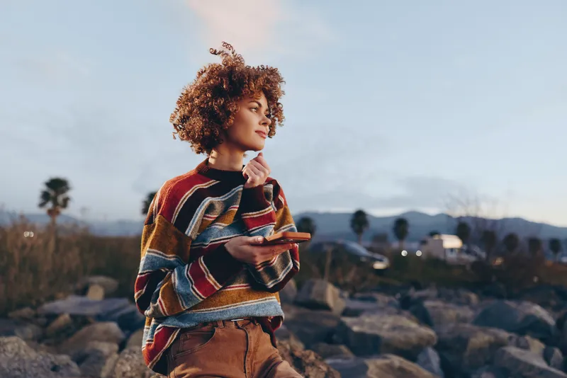 Woman lifestyle photo outdoors in mountains at sunset with curly hair wearing patterned sweater standing on rocky terrain thoughtful pose hand near chin soft evening light