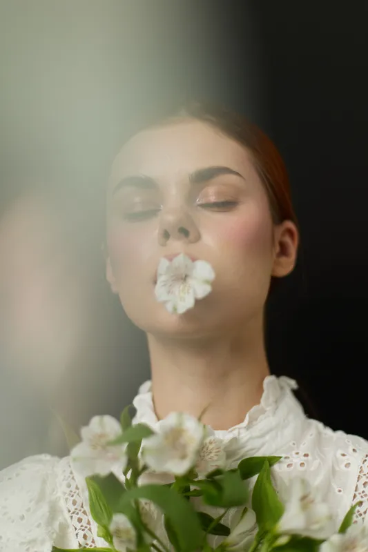 A serene young woman with a flower in her mouth, wearing a vintage white blouse, exuding calmness against a dark background, capturing the essence of natural beauty and tranquility