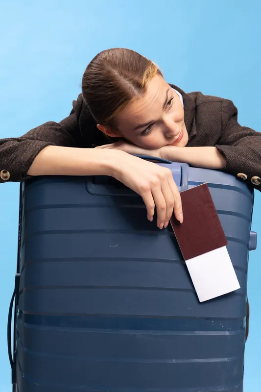 Young woman leaning on blue suitcase holding passport and ticket, looking thoughtful. Concept of travel planning, waiting, and vacation preparation with casual outfit on blue background.