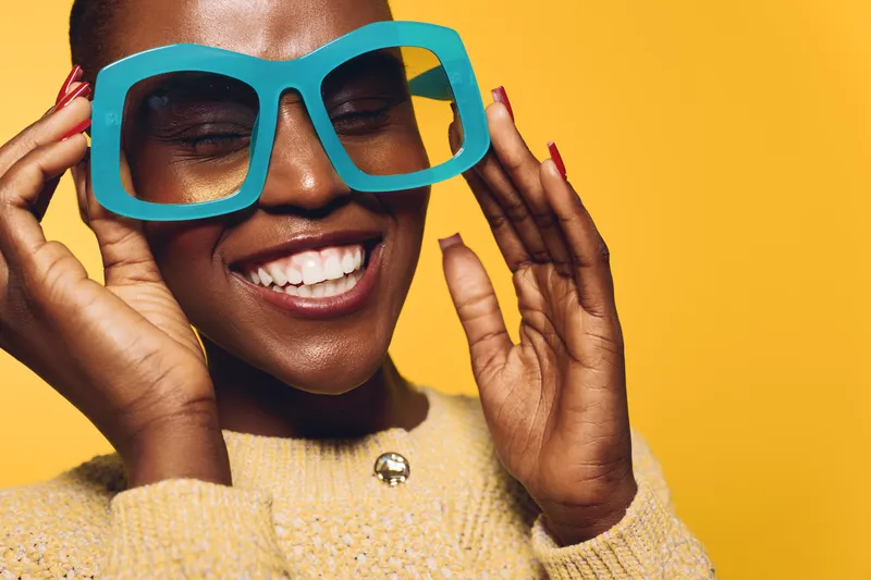 Happy smiling black woman with dark skin wearing large blue sunglasses and beige sweater poses against yellow background. Concept for fashion, joy, and lifestyle with natural beauty.