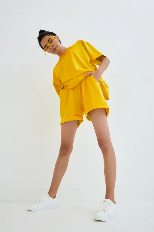 Young woman wearing bright yellow casual outfit and white sneakers, posing confidently against white background, showcasing modern streetwear fashion and summer style.