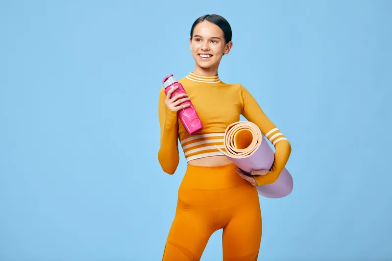 Sporty woman in orange athletic wear stands against a blue backdrop, holding a pink water bottle and a rolled yoga mat, ready for a fitness session and workout routine.