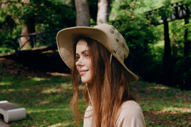 Woman hat portrait outdoor nature smile: Young woman wearing a wide brim hat in a park, side profile portrait with long hair, relaxed expression and soft natural light among green trees.