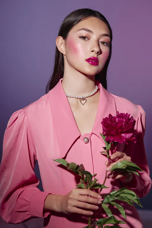 Young woman in a stylish pink dress with bold makeup poses holding a vivid flower. Studio portrait with creative beauty and fashion concept on purple background.