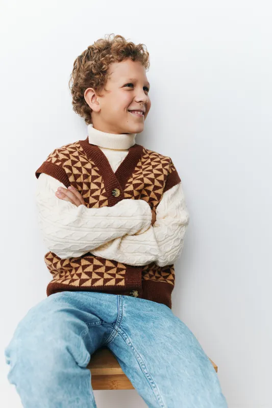 Authenticity captures a playful school lifestyle moment, featuring a cheerful child in a cozy knit vest, casual jeans, and a relaxed pose against a clean backdrop.