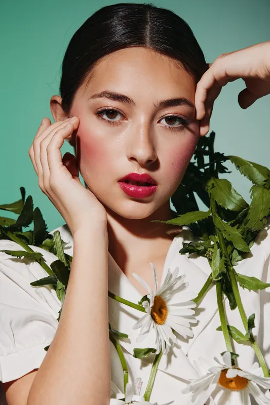 Young woman with radiant clear skin poses with delicate white daisies against a mint background. Her red lips and natural makeup highlight a fresh beauty fashion concept.