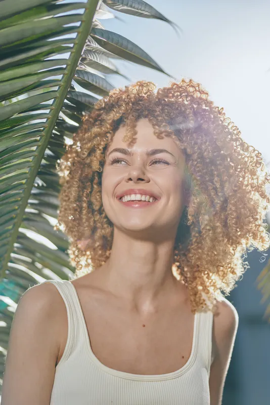 smiling woman with curly hair enjoys sunny tropical background, warm natural light enhancing bright joyful mood and relaxed summer atmosphere outdoors