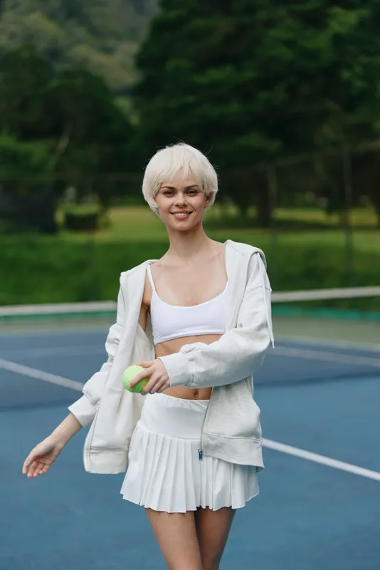 Young female tennis player smiling and walking on the court, dressed in white sportswear, showcasing a bright and vibrant atmosphere, fit lifestyle, and joy of sports