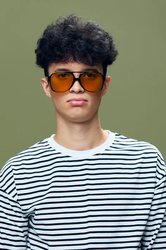 Young man wearing large orange sunglasses and striped shirt with curly hair looking indifferent on green background, casual style portrait with muted colors.