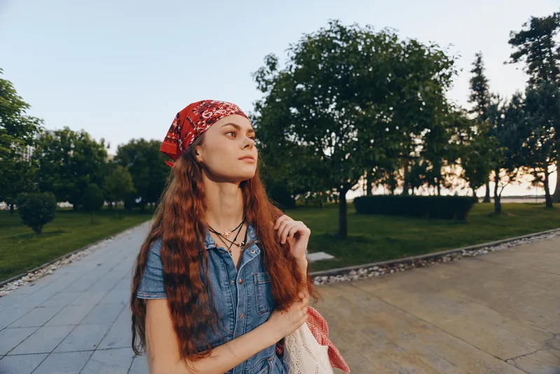 lifestyle woman with long hair in modern boho-western denim street style wearing red bandana, outdoors in park with film color tones and natural light, casual summer fashion