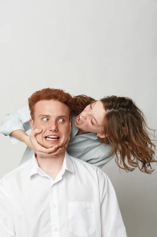 Playful young woman with curly hair teasing surprised redhead man in white shirt on light background. Concept of fun, friendship, humor, and casual interaction between friends or siblings.