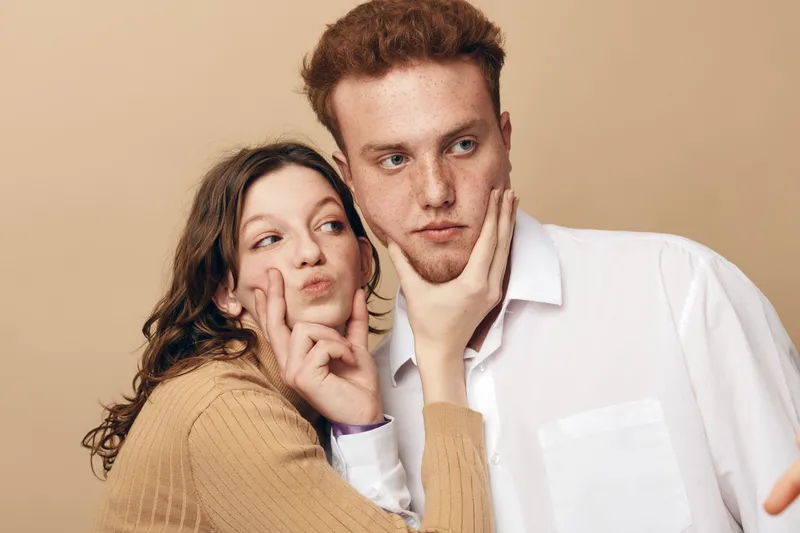 Young couple posing for selfie with playful expressions, woman making peace sign and man looking serious. Casual clothing and neutral background create relaxed, intimate mood.