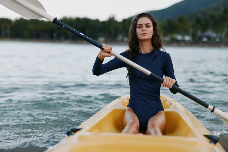 Young woman paddling a yellow kayak on calm water, wearing a dark blue wetsuit, focused expression as she rows toward the open lake and distant shore.