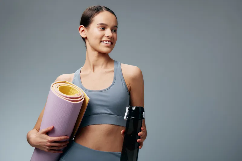 Female fitness enthusiast in sporty outfit holding a yoga mat and a water bottle, ready for a training session. Focus on fitness, sport, health and motivation in a clean studio.