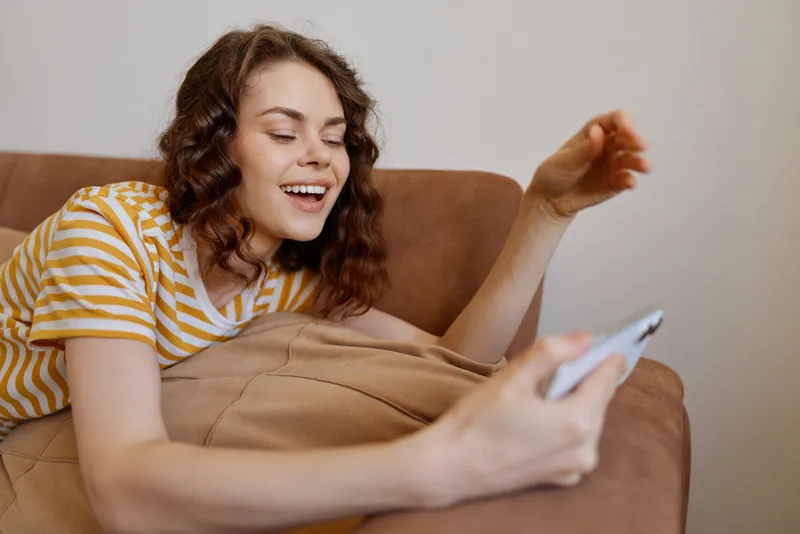 Happy woman relaxing at home, wearing a striped shirt, sitting on a couch and using a smartphone, warm natural light, cozy living room, relaxed mood, everyday lifestyle