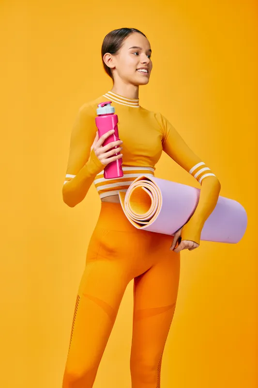 Fitness portrait of a athletic woman in bright orange outfit holding a pink water bottle and rolled yoga mat against a vibrant yellow backdrop, ready for training and motivation.