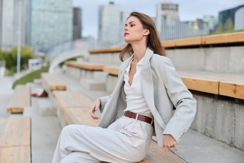 confident woman in business attire sitting outdoors on urban stairs with modern cityscape background, stylish beige suit and white shirt, professional and thoughtful pose