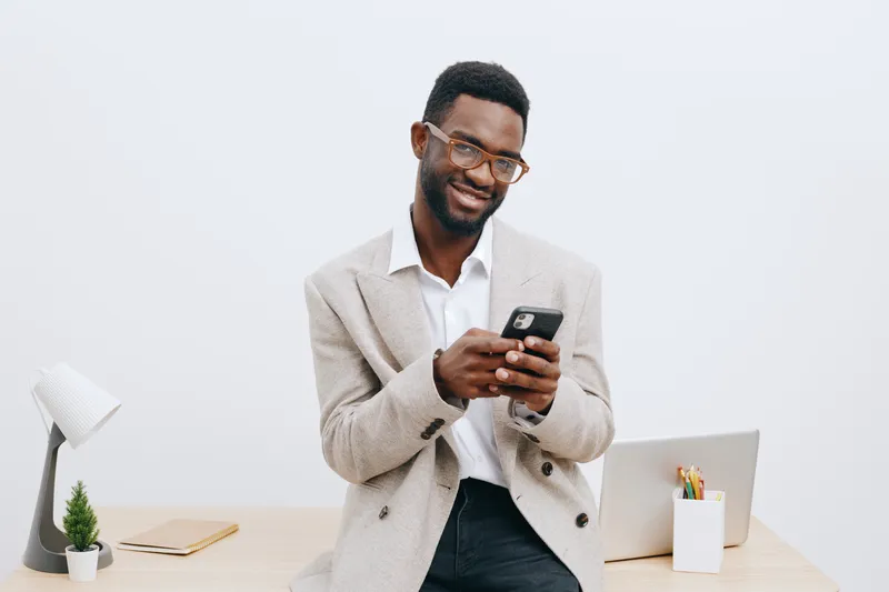 Smiling happy man with glasses in casual sweater using smartphone in modern office environment with minimalist decor and workspace accessories isolated on white background studio portrait. Business li