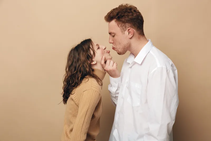 Young couple in casual clothes shares a tender moment, man gently holding woman’s chin as they lean in for a kiss. Intimate love and affection concept on beige background.