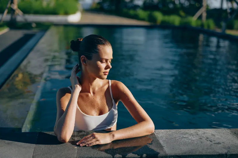 Relaxed woman rests at pool edge in a white sleeveless top, sunlit water shimmers, casual summer lifestyle, peaceful moment by the poolside, thoughtful mood.