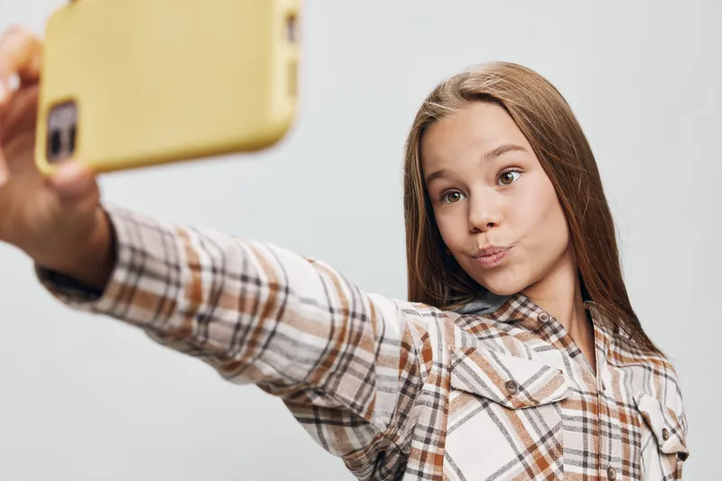 Teenage girl in plaid shirt taking selfie with smartphone against plain background. Young female, casual clothing and modern mobile phone for social media content or communication.