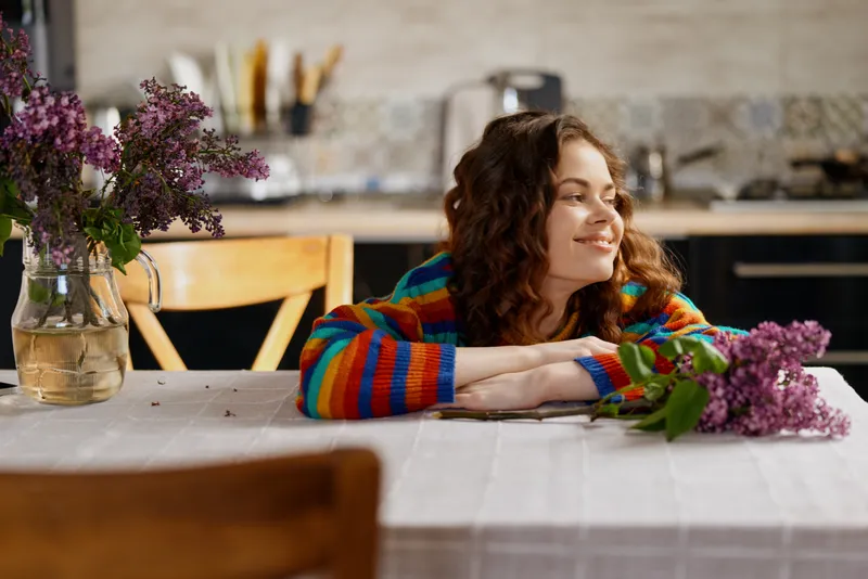 Young woman with curly hair rests her head on a dining table in a cozy home setting, smiling softly beside lilac flowers and warm natural light.