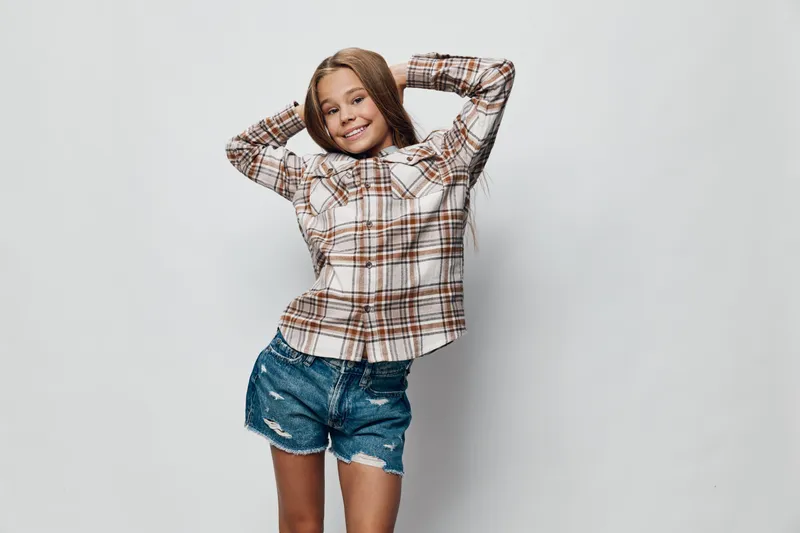 Smiling girl with long hair wearing a plaid shirt and denim shorts posing with hands behind head on white background in casual style. Happy child with relaxed posture.