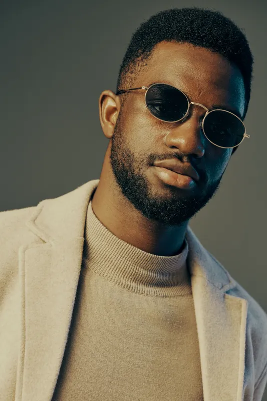 Stylish man wearing sunglasses and a beige coat, exuding confidence and trendiness in a modern urban setting with a cool color palette