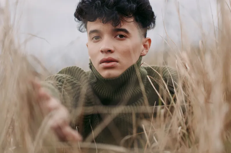 A thoughtful portrait in tall grass communicates authenticity through candid emotion and natural light, while a knitted sweater grounds the scene with honest, quiet presence.
