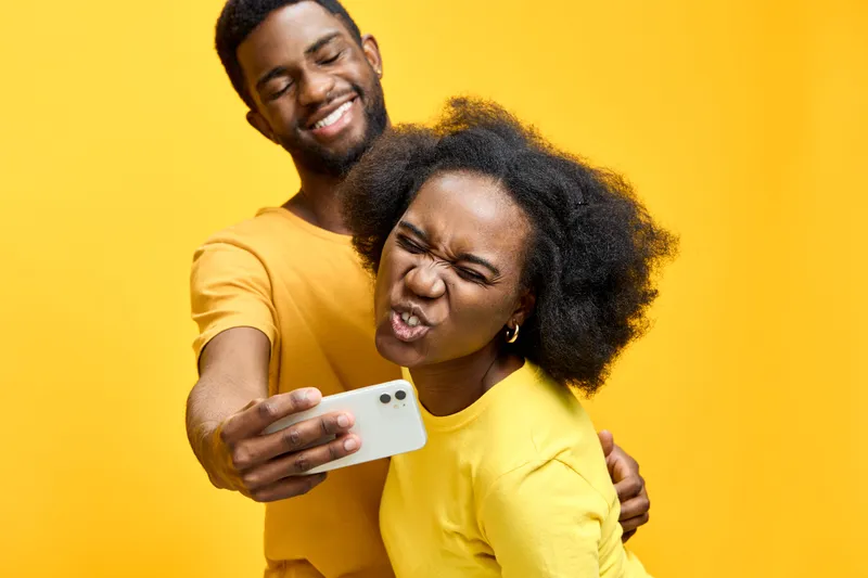 Happy couple taking selfies with a smartphone against a vibrant yellow background, showcasing joy and playful emotions in matching outfits for a cheerful lifestyle concept