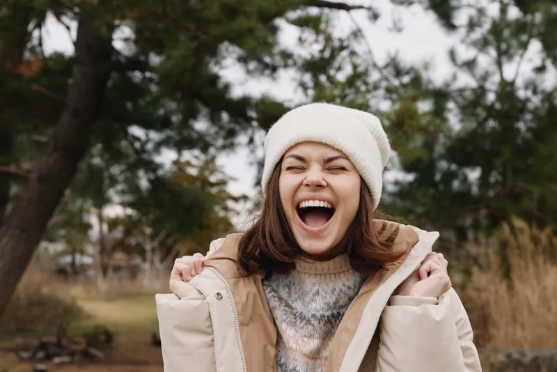 Woman in winter jacket outdoors, laughing with open mouth, wearing knit hat, a park scene around her, bright energy and candid happiness captured in a cool seasonal setting.