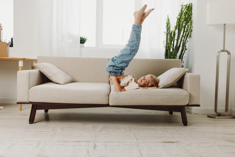 Happy child lying upside down on a beige sofa in a bright living room, playing with a book. Concept of childhood joy, home comfort, and playful learning.
