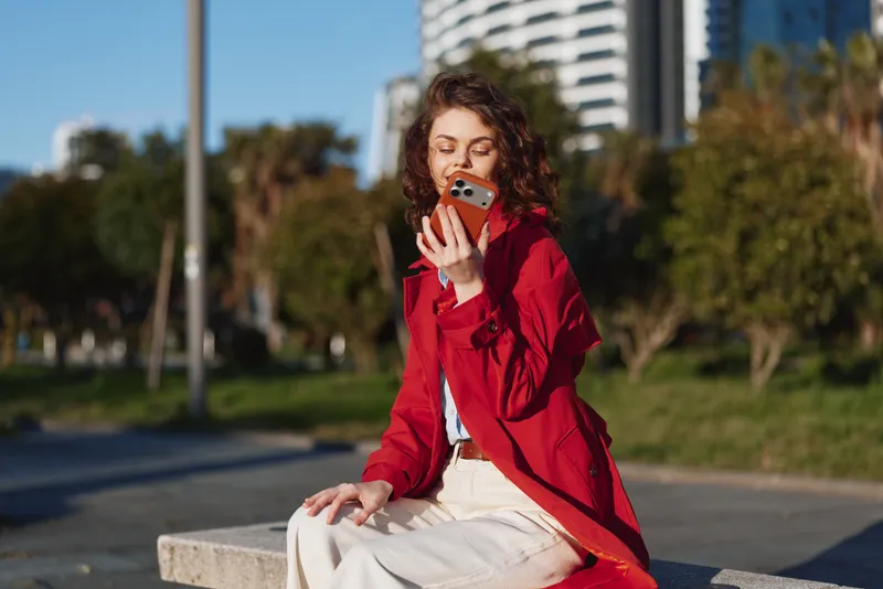 A young woman in a red jacket sits on a bench in a sunny outdoor park, focused on her smartphone, capturing a casual urban moment with a relaxed vibe.